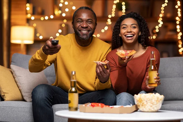 Emotional black couple watching TV together at home, eating pizza, drinking beer, laughing, copy space. Happy man and woman having fun together during winter holidays, love and relationships concept