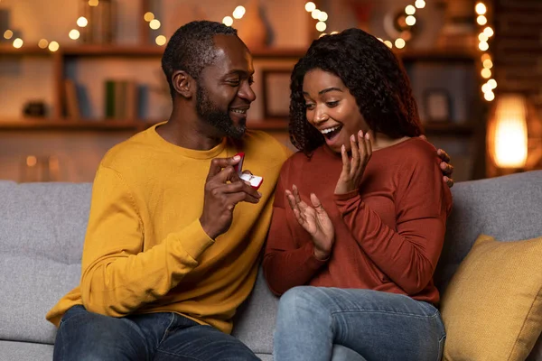 アフリカ系アメリカ人のハンサムな男を愛することは 照明で飾られたリビングルームの自宅でソファの上に婚約指輪で彼の驚くべきかなりのガールフレンドを驚かせました — ストック写真