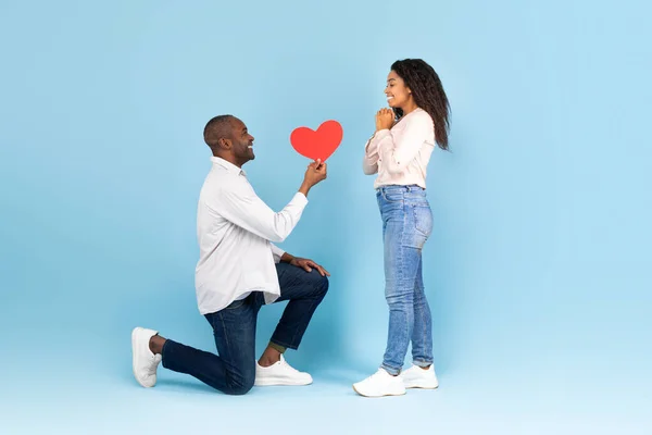 浪漫的非洲裔美国男人双膝跪着 把红心卡片送给兴奋的黑人女人 摆出一副蓝色画室的姿势 — 图库照片