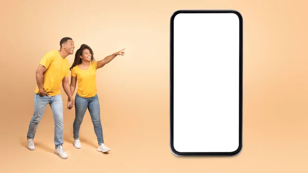 真棒的报价 兴奋的年轻黑人夫妇一边走一边指着屏幕空白的巨型手机 广告网站或应用程序 设计模型 横幅全景 黄色工作室墙 — 图库照片