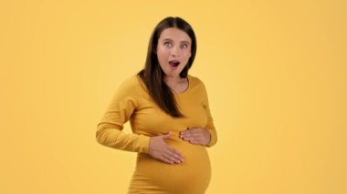 Baby activity. Young excited pregnant woman touching her belly, enjoying baby pushes and laughing, orange studio background, slow motion, empty space