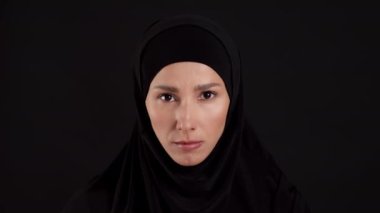 Save Iranian women. Close up portrait of young serious muslim woman wearing traditional hijab raising head and looking at camera, black studio background, slow motion