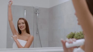 Personal hygiene habits. Young positive woman wrapped in towel applying antiperspirant on her armpits, looking at mirror at bathroom, slow motion, free space