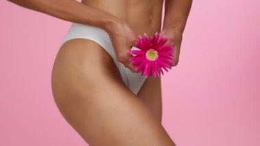 Female hygiene concept. Close up shot of unrecognizable young woman holding flower near her panties, pink studio background, slow motion