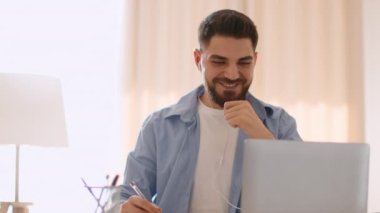 Convenient work. Young positive arab guy working on laptop and listening music online via earphones, working on laptop at home office, tracking shot, slow motion, empty space