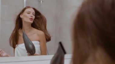 Bathroom fun. Young carefree caucasian lady wrapped in towel singing into hairdryer, drying her hair after shower, looking at mirror, tracking shot, slow motion, free space