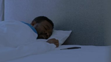 Problems with dream. Young african american man waking up in bed, looking at phone, checking time and keep sleeping late in night, tracking shot, slow motion, empty space