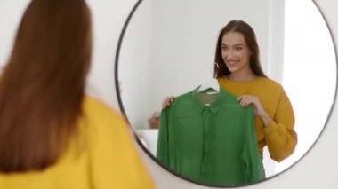 Young pretty lady choosing outfit for date, trying blouse on and smiling, preparing for business interview or date at home, looking at mirror, tracking shot, slow motion, free space