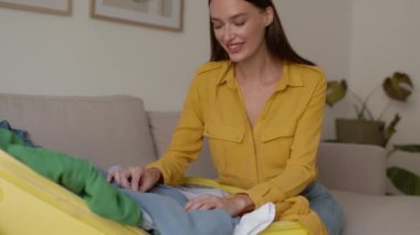 Young carefree woman tourist packing luggage for vacation, putting clothes into suitcase at home interior, tracking shot, slow motion, free space