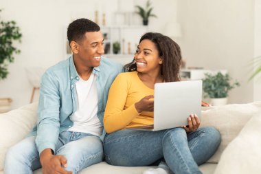 Smiling young african american lady show laptop to guy, recommend website and app for work and study, sitting on sofa in living room interior. Funny video in social networks, ad and offer at home
