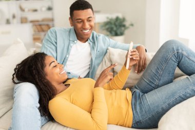 Smiling young black couple watch video on smartphone, enjoy rest and relax together in free time on sofa in living room interior. New app, chat in social network, lifestyle, ad and offer at home