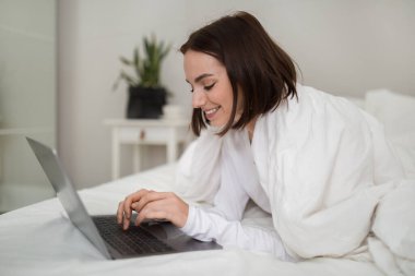 Relaxed happy beautiful brunette young woman in white pajamas chilling in bed with modern laptop, typing on computer keyboard and smiling, surfing on Internet, chatting on dating website, copy space