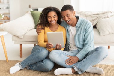 Happy laughing young african american couple hugging looking at tablet webcam, watch funny video, enjoy spare time in living room interior. Video call, meeting, chat and new app at home due new normal