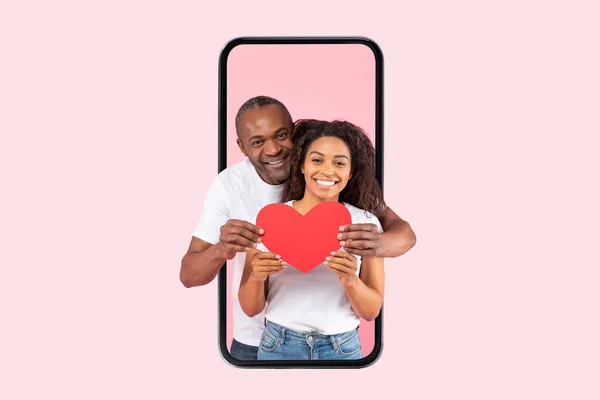 爱和关心 快乐的非洲裔美国配偶拥抱着红纸卡片的心脏 站在智能手机屏幕上 背景是粉色的工作室 圣瓦伦丁日 — 图库照片