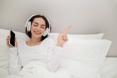 Tranquill emotional young brunette lady wearing white pajamas listening to music, singing karaoke and dancing in bed at home, using smartphone with mockup and newest wireless headphones, copy space