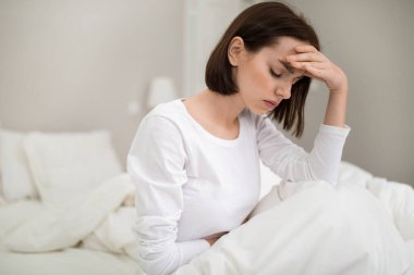 Young sick brunette woman with headache sitting in bed with closed eyes and touching her forehead at home, unhappy lady suffering from migraine or hangover, copy space