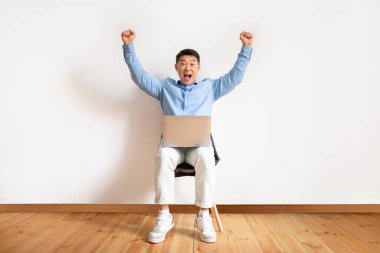 Overjoyed asian mature man making YES gesture, sitting on chair with laptop, celebrating success or achievement against white studio wall, full length. Male feeling excited over big win