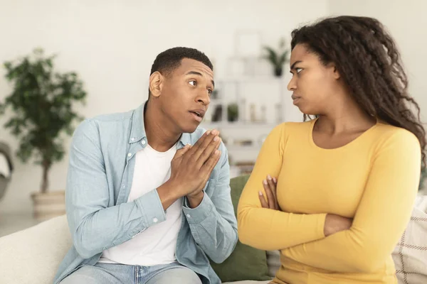 Sad young african american man asking for forgiveness from angry offended female in living room interior, free space. Apologize, sorry, relationship problems, people emotions after quarrel at home