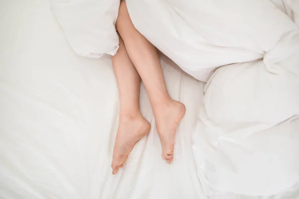 Lying on bed female legs under white blanket. Legs of young woman, that lies on her bed in bedroom under the eiderdown. Unrecognizable lady sleeping with feet out of duvet, cropped, top view