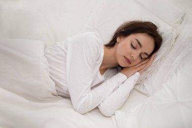 Good sleep for good health concept. Beautiful brunette young woman in white pajamas resting in white bed at home, high angle view of peaceful lady sleeping in her comfortable bed