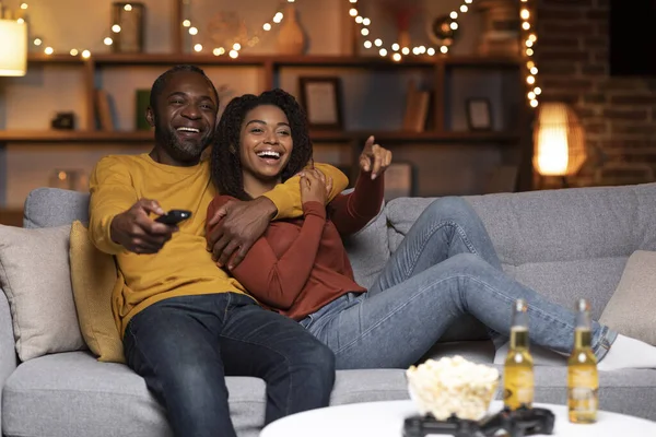 Joyful african american couple watching TV together at home, laughing, bonding, copy space. Happy black man and woman having fun together during winter holidays, love and relationships concept