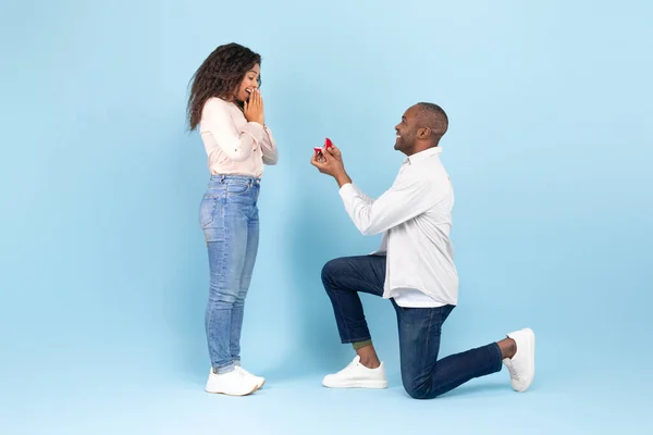可爱的黑人男子单膝站立 并提供订婚戒指他心爱的年轻女子蓝色工作室背景 侧视图 中年男子向情人求婚 — 图库照片