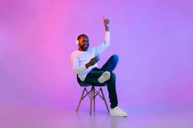 Cool mature black man in casual sitting on chair over neon light studio background, listening to music and moving his hands, using phone and wireless headphones, enjoying stereo sound, copy space