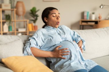 Pregnancy pain. African american pregnant woman touching belly while sitting on couch at home, lady suffering painful contractions and making breathing exercises to relief ache clipart