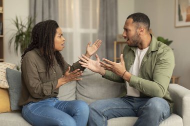 Emotional african american lovers having quarrel, sitting on sofa in front of each other, yelling and gesturing, experiencing difficulties in marriage