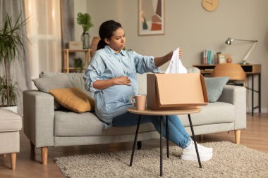 Discontented young pregnant black woman unpacking box and holding delivered clothes, sitting on sofa at home, free space. Delivery service problems concept