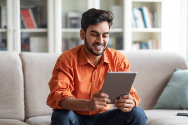 Happy stylish handsome young middle eastern guy sitting on couch in living room, using digital tablet at home and smiling, chatting with friends, websurfing, using nice mobile app, copy space