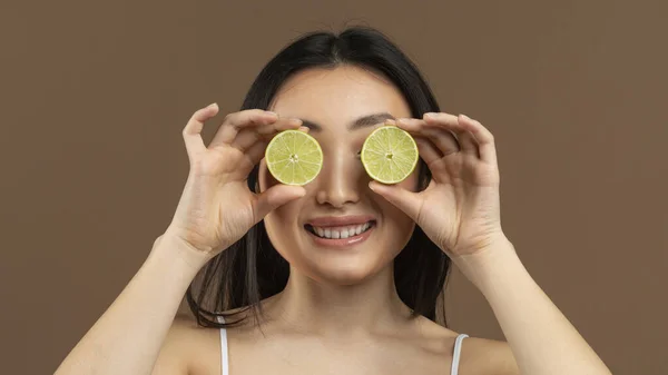 Vitamins for beauty and skin care. Asian lady covering eyes with lime halves, standing over brown studio background, panorama. Young korean woman holding citrus fruits near her face