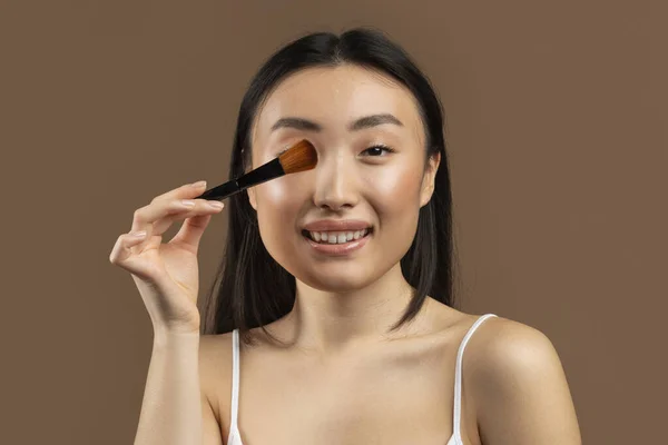 Beautiful happy asian lady holding makeup brush, applying blush on face and smiling at camera, japanese woman powdering her cheeks, posing on brown background