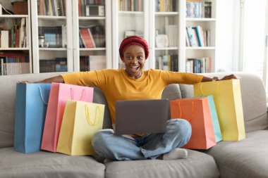 Happy excited pretty young black woman in red turban shopaholic sitting on couch with lot of colorful shopping bags, using laptop at home. Retail, e-commerce, online shopping concept, free space
