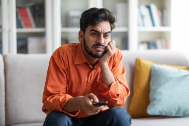 Bored unhappy handsome middle eastern millennial guy in orange shirt sitting on couch at home, switching tv, looking for nice film or TV show, free space. Domestic entertainment concept