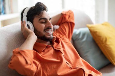 Joyful happy handsome stylish middle eastern millennial guy in casual reclining on sofa in living room, using wireless headphones, singing songs at home, karaoke mobile app, copy space, closeup shot