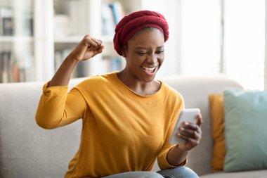 Emotional happy african american young woman wearing traditional red turban using smartphone at home, sitting on couch in cozy living room, looking at gadget screen, gesturing, good news, free space