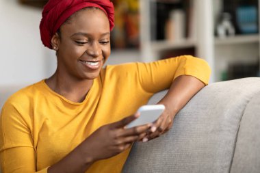 Positive cheerful relaxed pretty millennial black woman wearing red turban sitting on couch at home, using modern smartphone, chatting with guys on dating mobile app, copy space, closeup