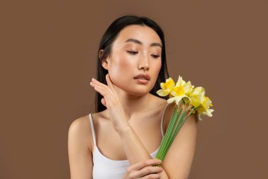 Young beautiful japanese lady posing with yellow daffodil flowers isolated on brown background, studio shot. Beauty and skin care, anti-aging treatment, spa procedure, ad and offer