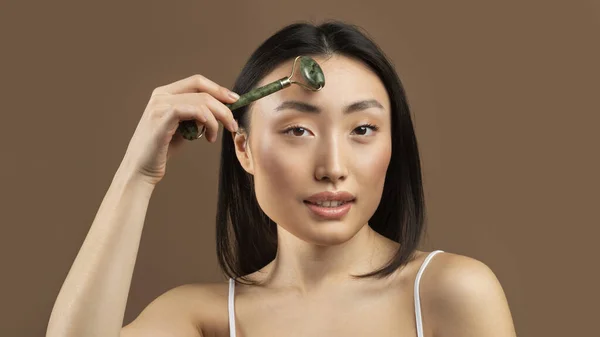 Beautiful japanese woman making lymphatic drainage facial self massage with jade roller on brown studio background, panorama. Young korean lady making rejuvenation skincare procedure