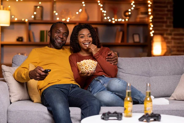 Happy cheerful relaxed black spouses handsome middle aged man and young woman in casual sitting on couch, watching TV at home decorated with lights, drinking beer, eating popcorn, copy space