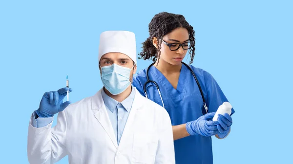 青いスタジオの背景で働く多人種間の医療チーム 白いコートの男と女と薬で注射器や瓶を保持する保護医療面の医師 パノラマ — ストック写真