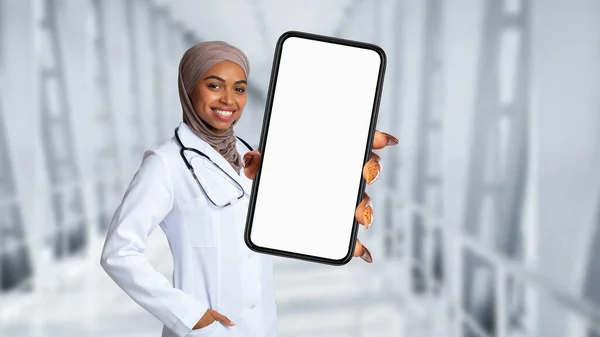 Black Muslim Doctor Lady Wearing Uniform Showing Big Blank Smartphone At Camera While Standing Over Clinic Hall Background, Young African American Female Physician Advertising Medical Offer, Mockup
