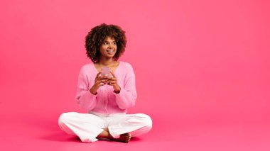 Glad inspired millennial african american curly lady with braces sit on floor, typing on phone, look at copy space isolated on pink background, studio. App for study, fashion blog, sale, ad and offer