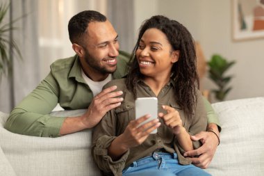 Loving young black spouses using cellphone together, resting on couch in living room, enjoying time at home, copy space. Happy african american couple checking new shopping application