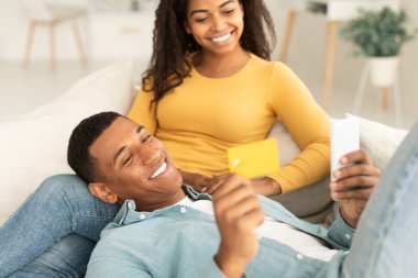 Happy young african american guy show smartphone and credit card to lady, shopping online together in spare time on sofa in living room interior. Financial app, banking, sale for shopaholics at home