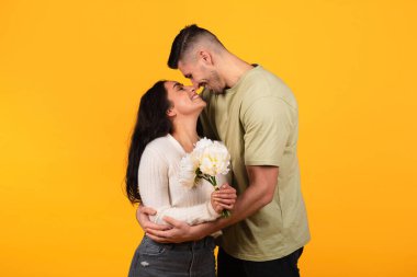 Happy millennial arabic guy hugging lady, gives bouquet of flowers, isolated on orange background, studio, free space. Couple enjoy love, romance and relationship, celebrate anniversary, date, holiday