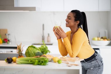 Smiling young african american female drinking glass of smoothie at table with organic vegetables, enjoy diet in modern kitchen interior. Cooking at home, health care, vegetarian food for weight loss