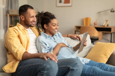 Excited african american pregnant couple relaxing with digital tablet on sofa at home, black spouses choosing and purchasing clothes for baby in internet
