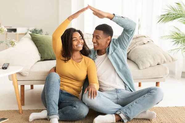 Cheerful millennial african american family make roof sign with hands, sit on floor in living room interior. Own house, credit and mortgage on real estate, dream for future, relationship and lifestyle